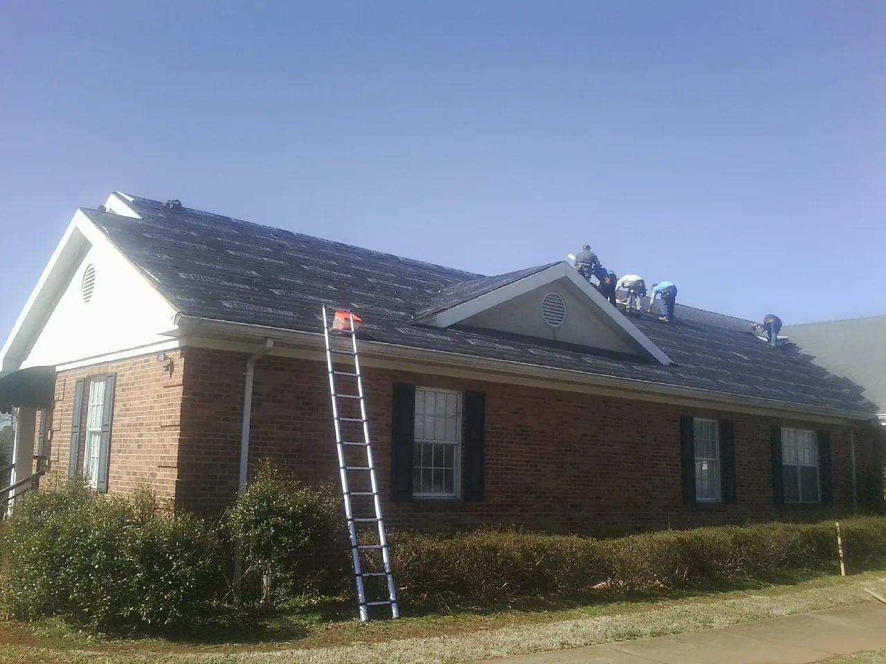 Crew tearing shingles off a brick ranch roof with a ladder set — Emergency Roof Repair work by our crew in Indio