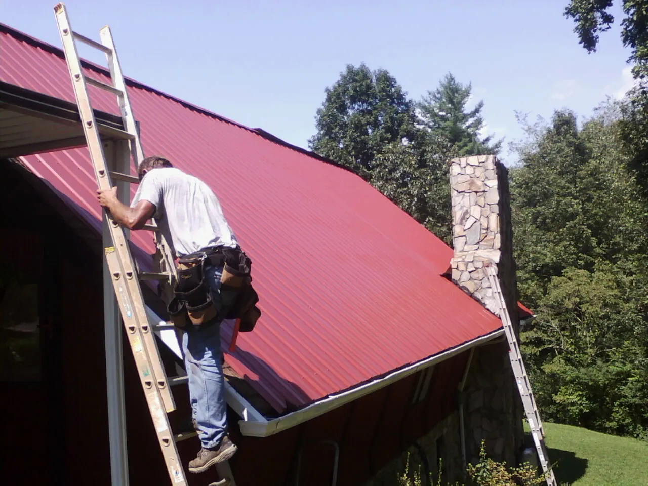 Roofer on a ladder beside a red metal roof and stone chimney — Soffit & Fascia Repair by Roofing Brothers in Indio